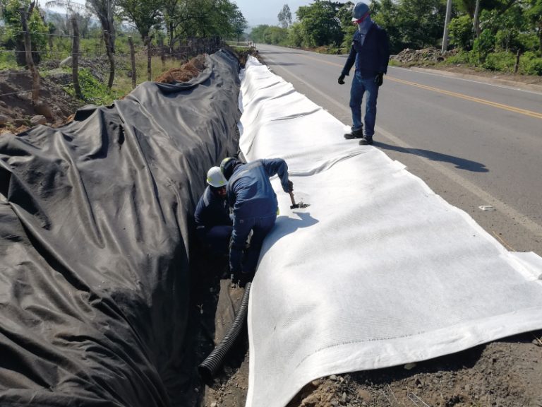 Operarios instalan un geodrén con tubería corrugada en una zanja al borde de una carretera