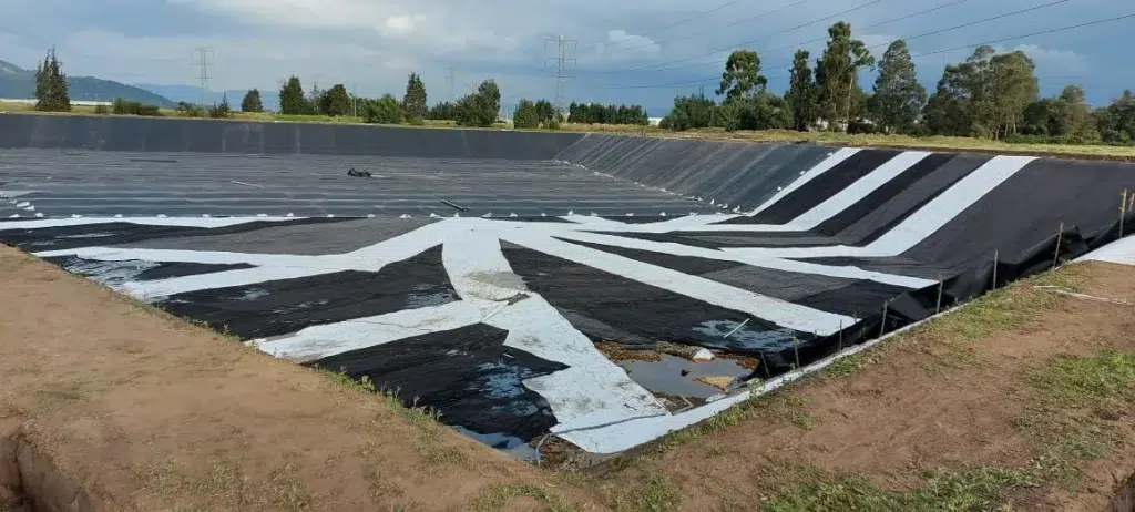 Piscina en construcción revestida con geomembrana negra y franjas blancas de geocompuesto sobre el fondo y los taludes