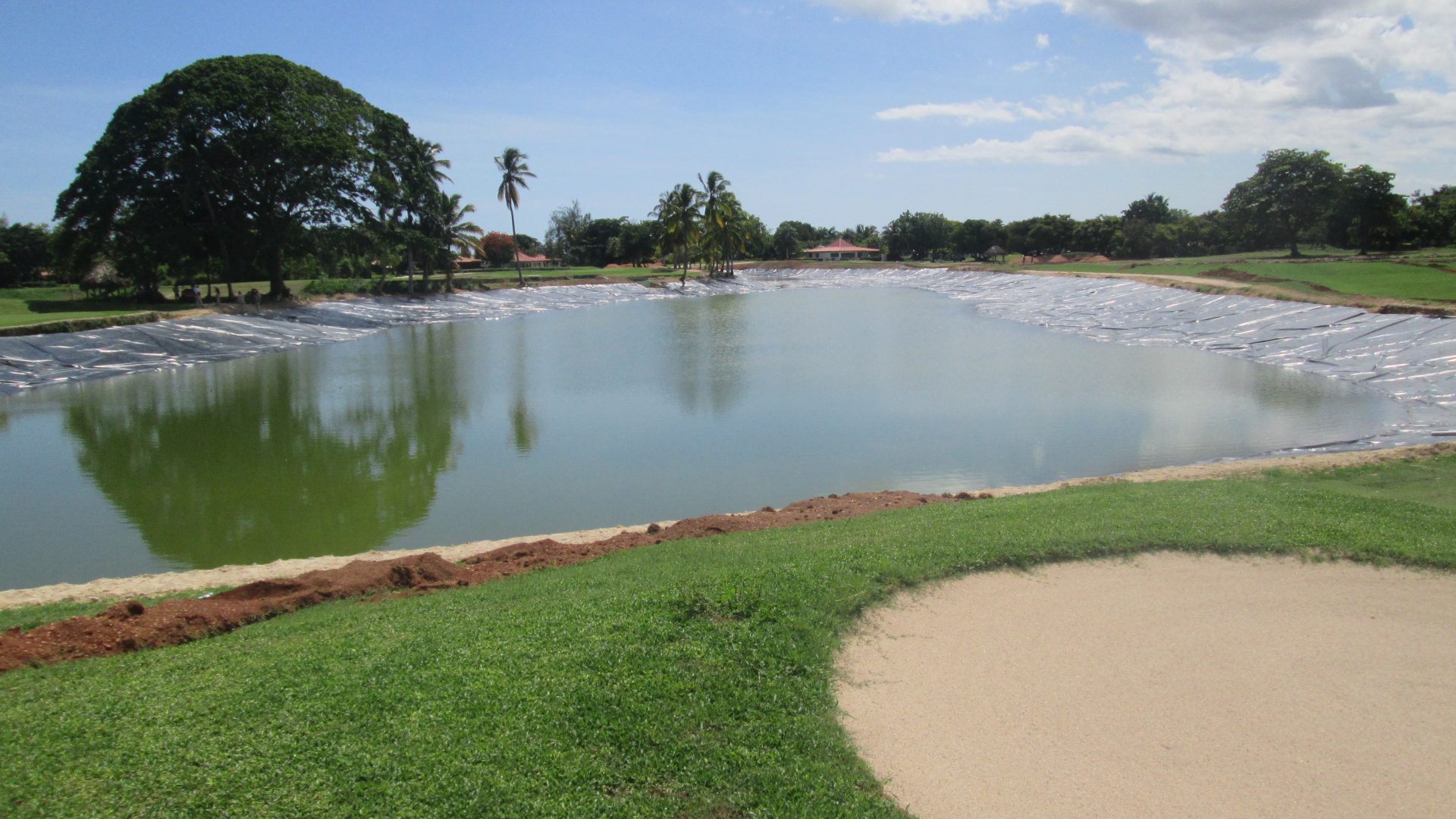 Estanque o laguna artificial en un campo de golf, parcialmente lleno, con el fondo revestido con una geomembrana negra.