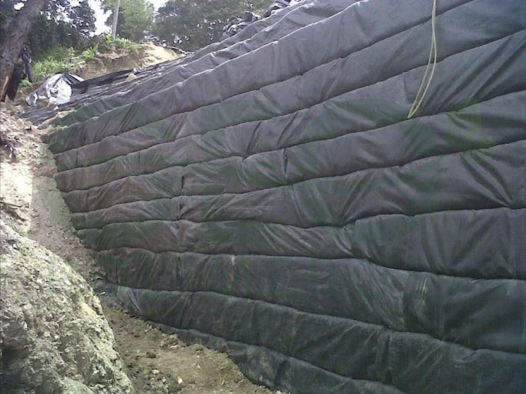 Muro en suelo reforzado con capas envueltas en geotextil negro, junto a un corte de terreno